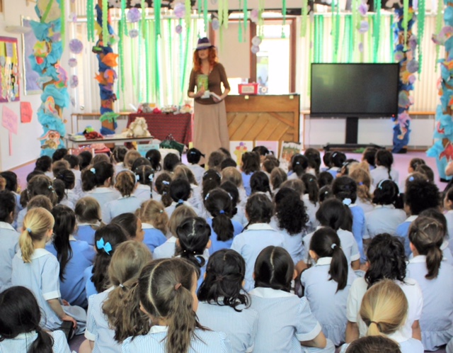 A class full of children, seen from behind, sat on the floor and looking up as Alison Grunwald, author, speaks to them holding her book